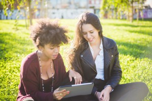 Bestie2 two multiethnic beautiful young woman black and caucasian using tablet sitting in a city park, lookind down the screen smiling - technology, social network, happiness concept