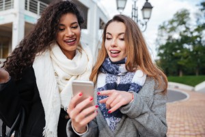 Happy two girlfriends looking on a smartphone and pointing at screen surprised. Caucasian girl wearing scarf in a cage print. African lady wearing burgundy scarf.