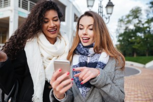 Bestie4 Happy two girlfriends looking on a smartphone and pointing at screen surprised. Caucasian girl wearing scarf in a cage print. African lady wearing burgundy scarf.